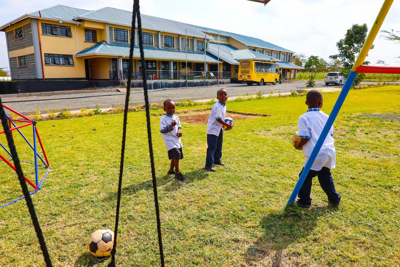 Young pupils playing football on the school field with the Kare Educational Centre buildings behind